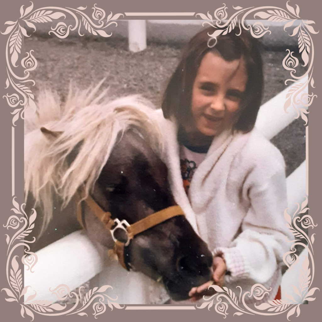 Image: A photo of Carrie as a child wearing a white sweater with chin-length brown hair and a round barrette. She is smiling but missing her front teeth and standing next to a pony that is nibbling her hand. 