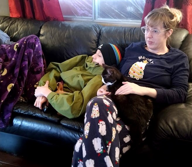 Image of Carrie looking exhausted with no makeup and a lazy bun in her hair, on a couch win her holiday jammies on. In her lap is Myrtle,  a black and white cat, and her son laying against her. They are looking in the distance at the TV.